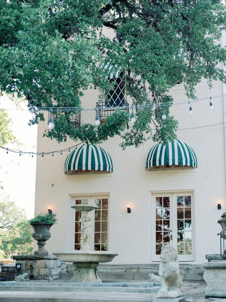 The side of the Laguna Gloria with green and white stripe shades over the windows and a neoclassical fountain.