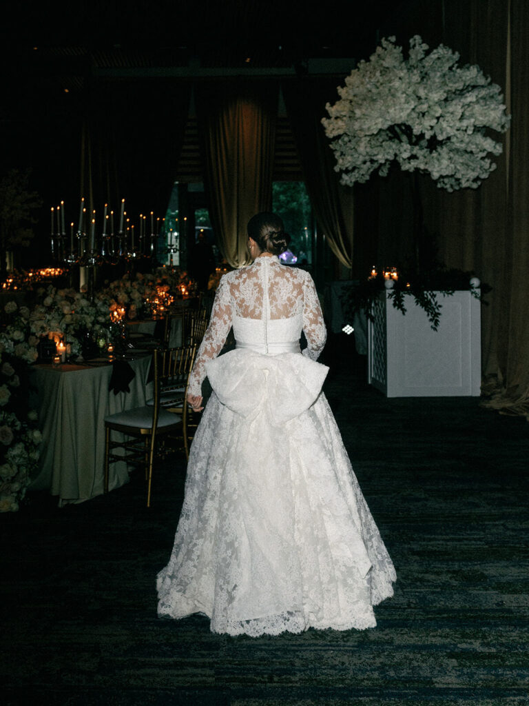A bride walks away into her reception wearing a long sleeve lace gown with a large bow at her waist for her Witte Museum wedding.