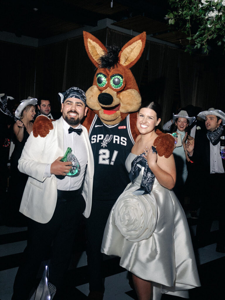 The bride and groom pose together with the mascot of the San Antonio Spurs basketball team, a tall coyote wearing a basketball jersey.