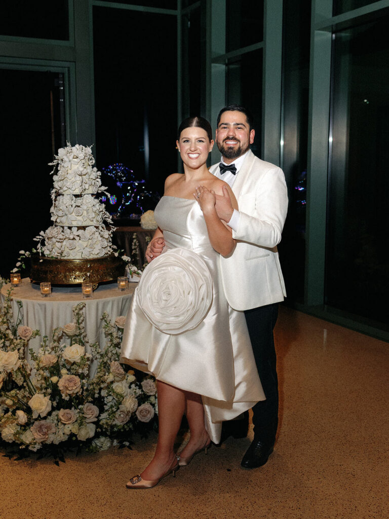 The bride and groom pose in their new reception looks, the bride in a knee-length satin gown with a large floral detail and the groom in a white tuxedo jacket for their Witte Museum wedding.
