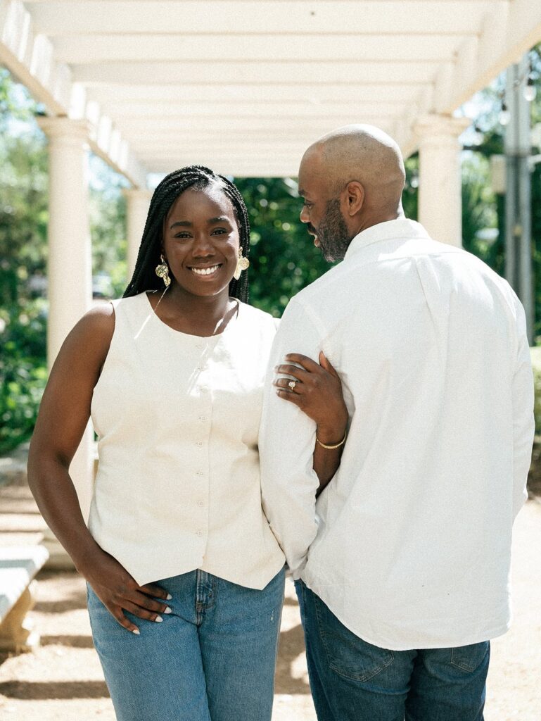A woman smiles and faces the camera with her arm linked in her fiance's who is turned away from the camera.