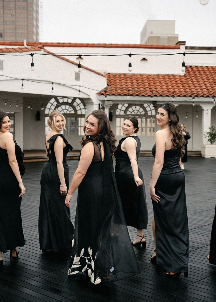 Bridesmaids in black dresses walk away from the camera but turn back and smile.