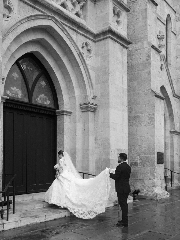 The groom holds the train of his bride's gown on the steps of the cathedral during their Witte Museum wedding.